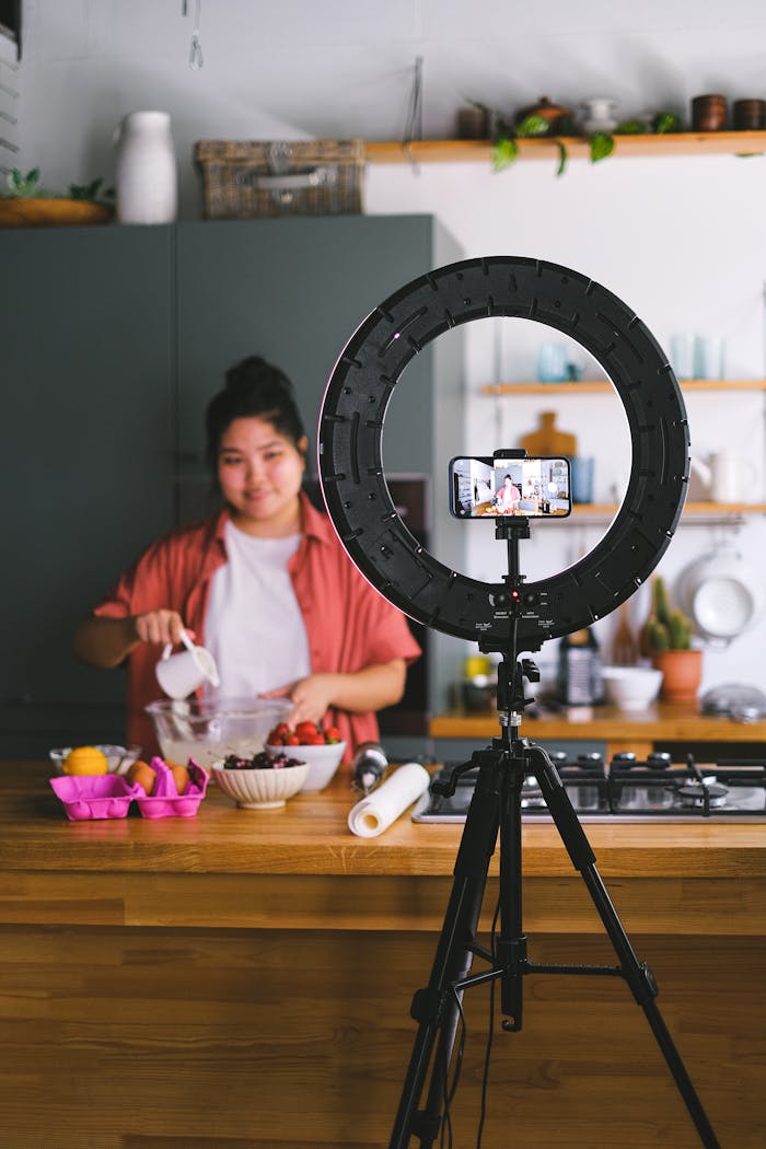 A woman prepares ingredients while filming a cooking vlog in a modern kitchen using a smartphone and ring light.